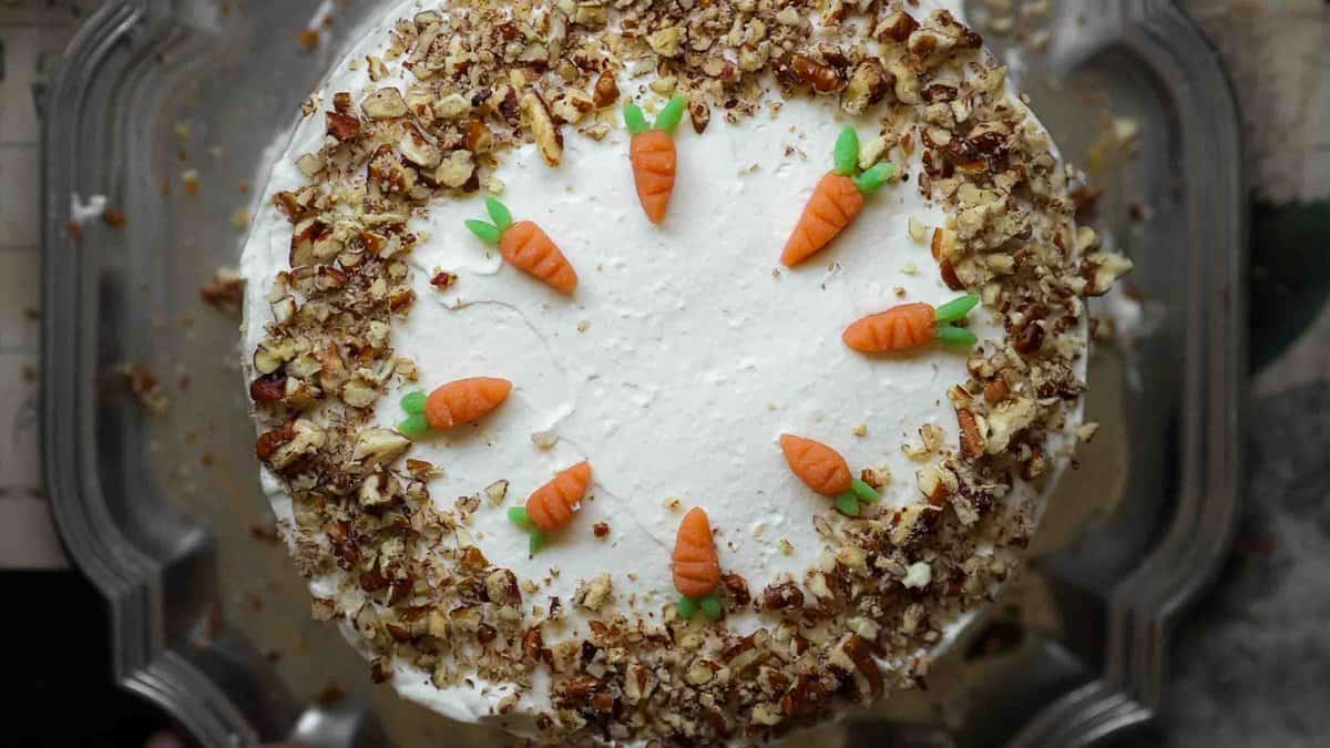 A round vegan carrot cake topped with white frosting, adorned with small fondant carrots in a circle on top. Chopped nuts line the edges, adding texture and contrast to the smooth icing.